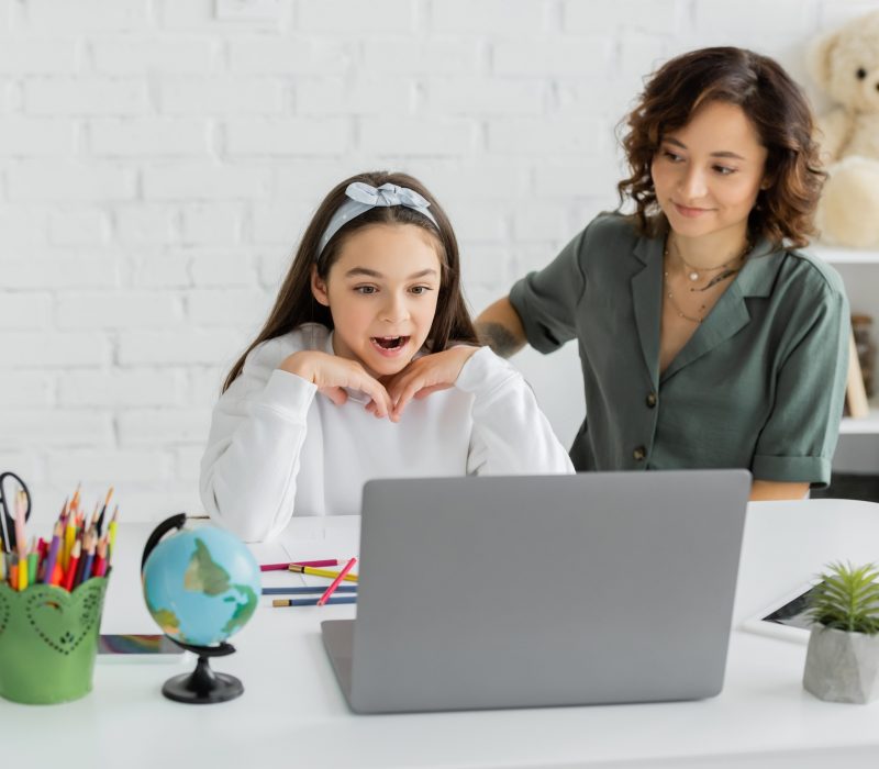 Preteen girl looking at laptop during speech therapy online lesson near mother at home